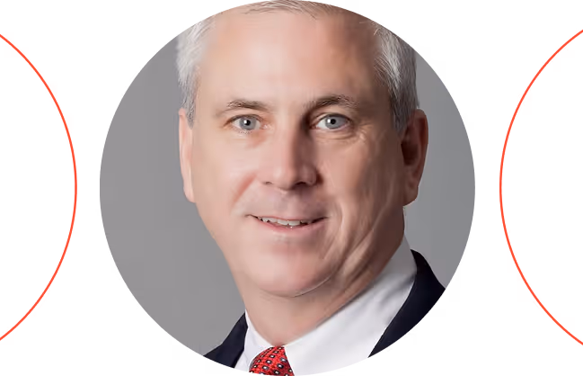 Headshot of a middle-aged man with gray hair, wearing a dark suit, white shirt, and red patterned tie, smiling against a gray background with red accents.