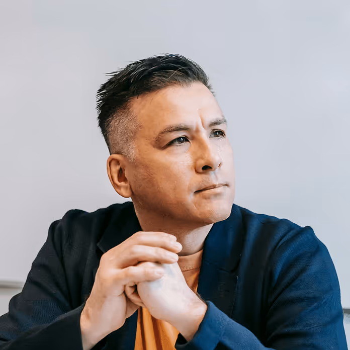 Man with short dark hair in a dark jacket and orange shirt sitting with hands clasped, looking thoughtfully to the side.