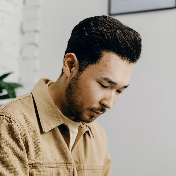Man with short dark hair and beard wearing a beige jacket looking downward thoughtfully.