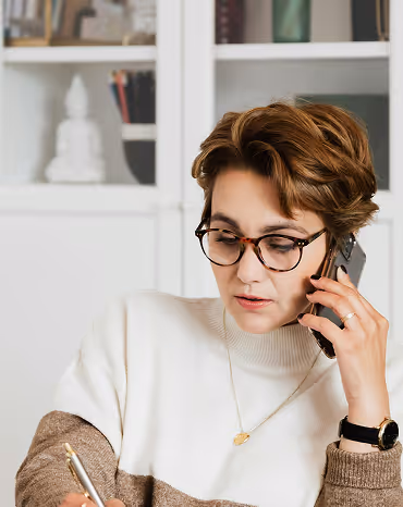 Woman wearing glasses talking on a phone while writing with a pen indoors.