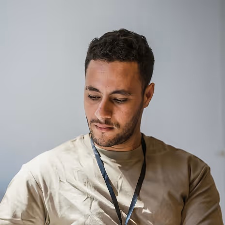 Man with short curly hair and a light beard wearing a beige shirt and a black lanyard, looking down.
