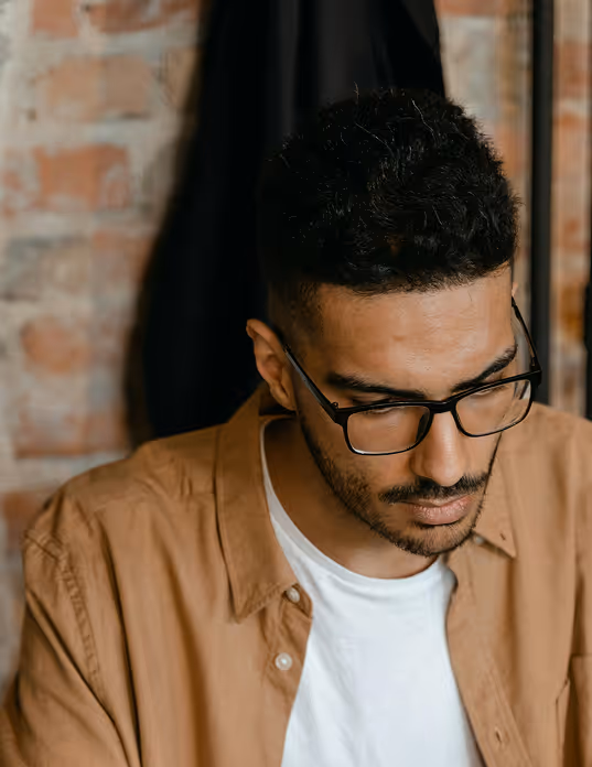 Man with short curly hair and glasses wearing a tan shirt over a white t-shirt looking down.
