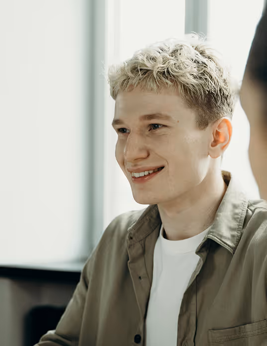 Young man with curly blonde hair smiling indoors wearing a white shirt and olive-green jacket.