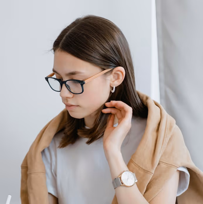 Young woman with brown hair wearing glasses, pearl earrings, a light gray shirt, and a tan jacket draped over her shoulders, looking down while touching her hair.