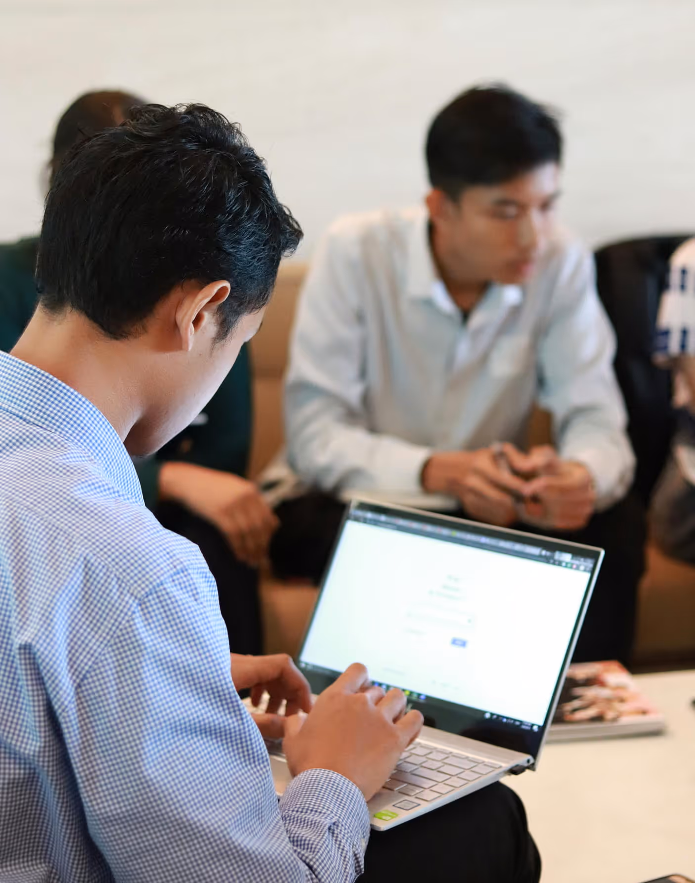 Man in a checkered shirt typing on a laptop with two others conversing in the background.