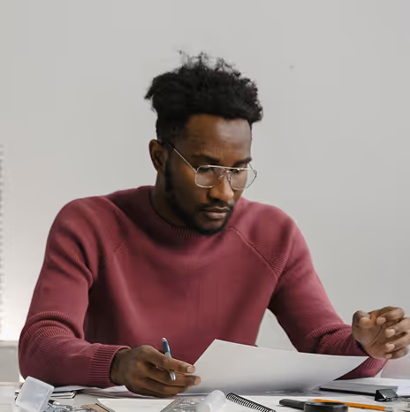Man with glasses in a maroon sweater reviewing documents at a desk with office supplies.