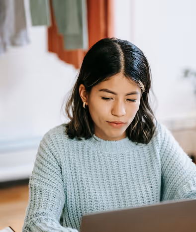 Young woman with black hair wearing a light gray sweater, focused on a laptop in front of her indoors.