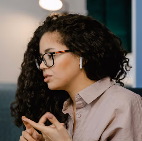 Woman with curly hair, glasses, and wireless earbuds talking and gesturing with her hands indoors.