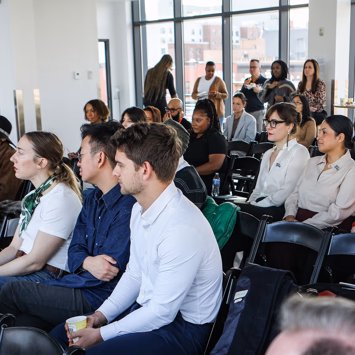 Diverse group of people sitting and standing in a bright room with large windows during an event.