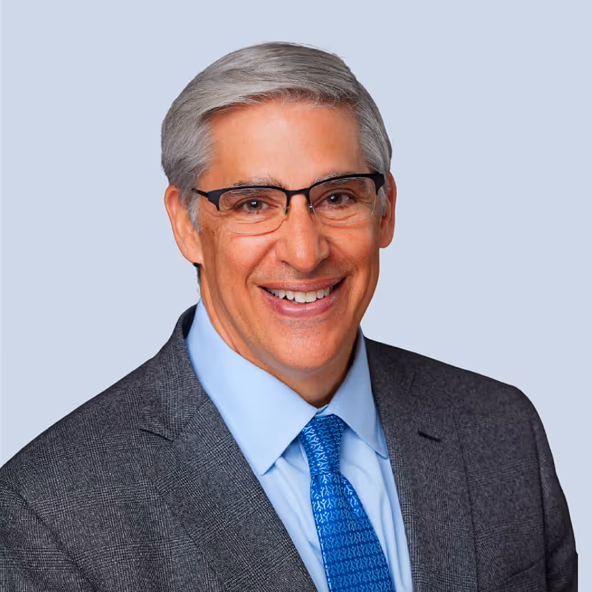 Smiling older man with gray hair, glasses, wearing a gray suit jacket, light blue shirt, and blue patterned tie on a light blue background.