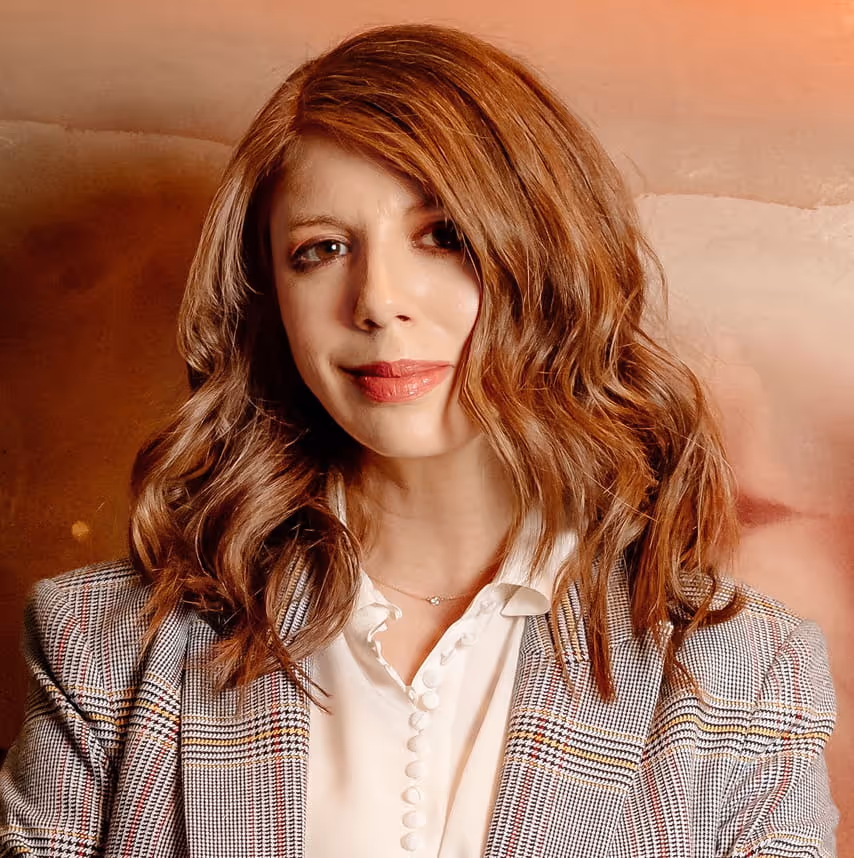 Woman with shoulder-length wavy auburn hair wearing a checkered blazer and white buttoned shirt, smiling softly against a warm brown and peach background.