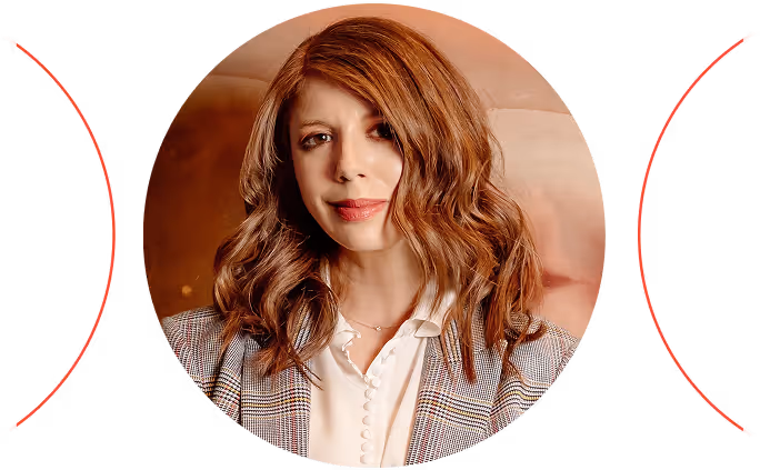Woman with wavy reddish-brown hair wearing a checkered blazer and white blouse, seated indoors.