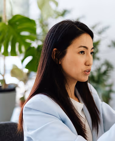 Young woman with long dark hair speaking, dressed in a light blue blazer, indoors with green plants in the background.