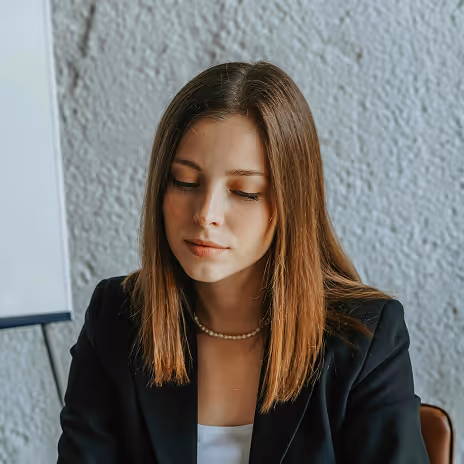 Young woman with straight brown hair wearing a black blazer and pearl necklace, looking down thoughtfully against a textured gray wall.