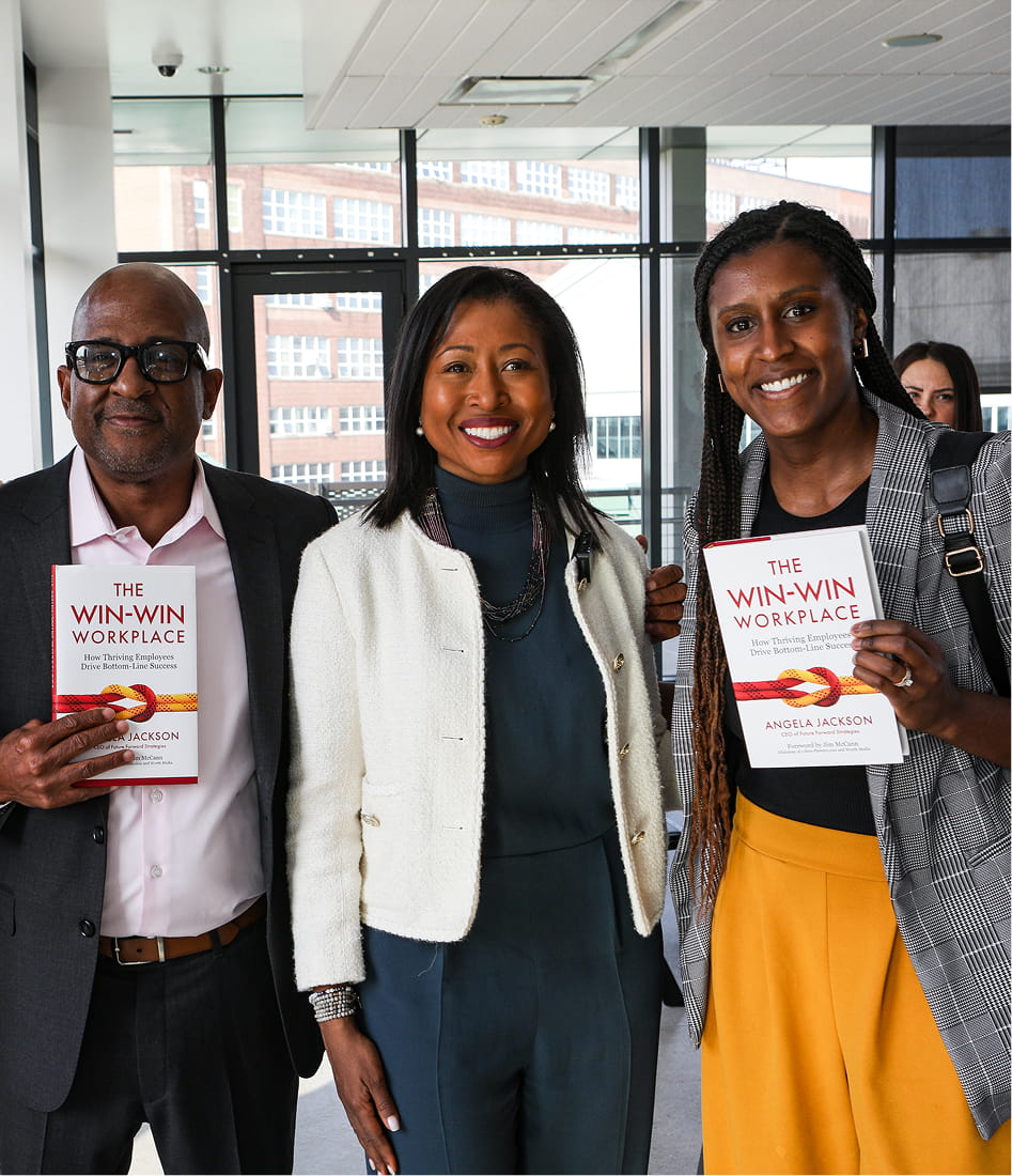 Three smiling people indoors, two holding copies of the book 'The Win-Win Workplace' by Angela Jackson.