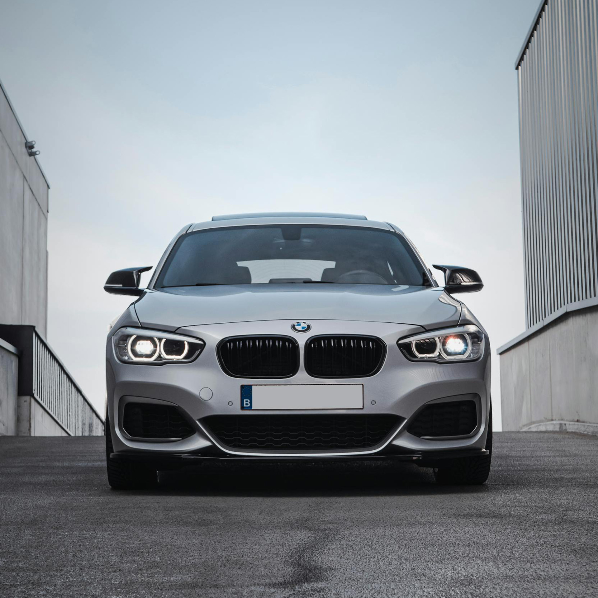 Front view of a silver BMW car with headlights on parked between two buildings under a cloudy sky.