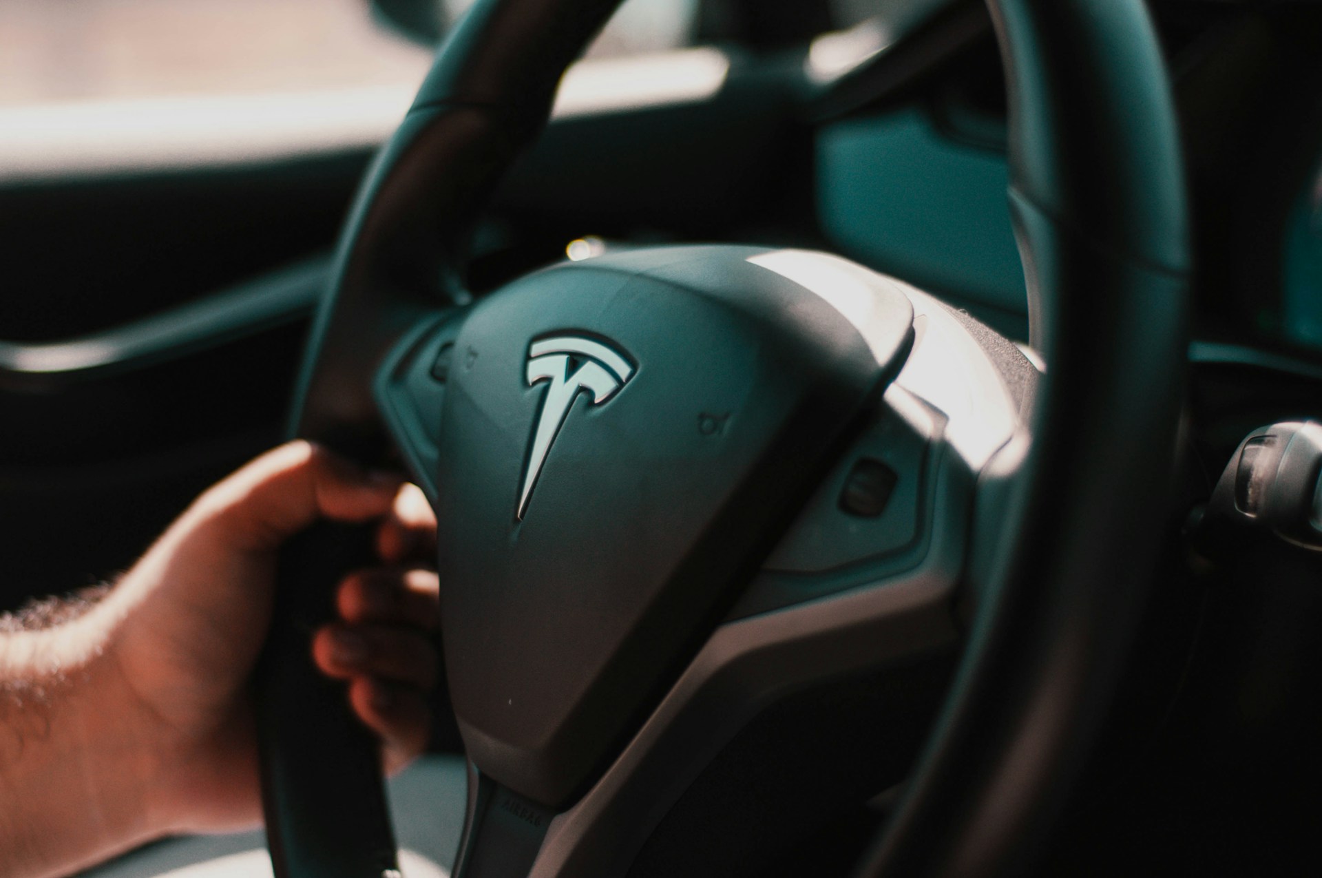 Close-up of a person’s hand on a Tesla car steering wheel with the Tesla logo visible.