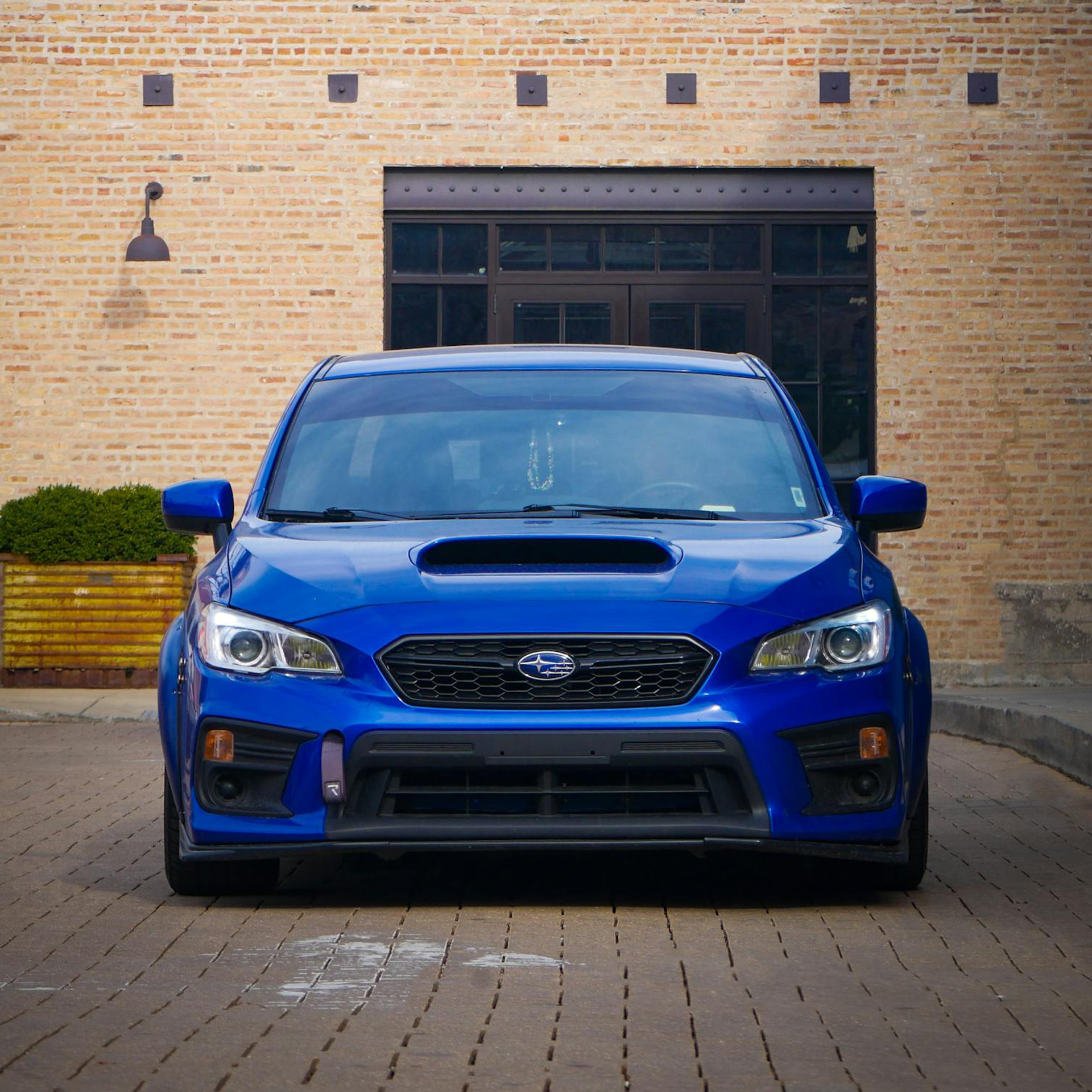 Front view of a blue Subaru car parked on a brick-paved street with a brick building in the background.