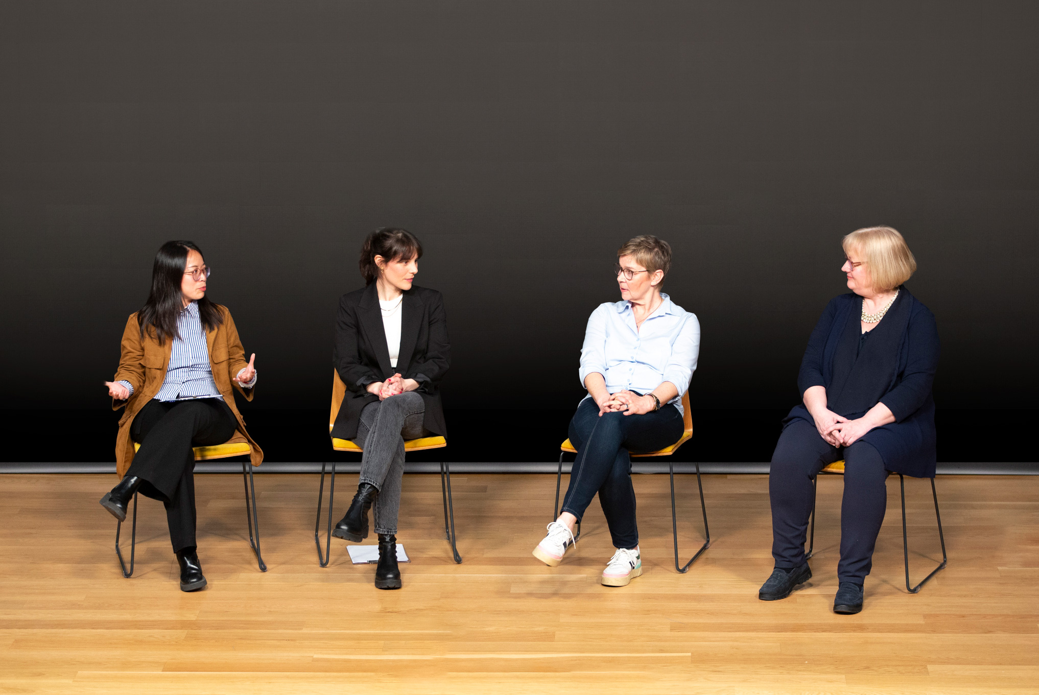 Four women seated on stage chairs, engaged in a discussion against a dark backdrop.