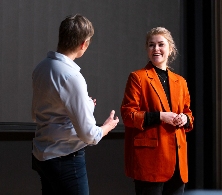 Two people engaged in conversation, one wearing an orange blazer smiling at the other.