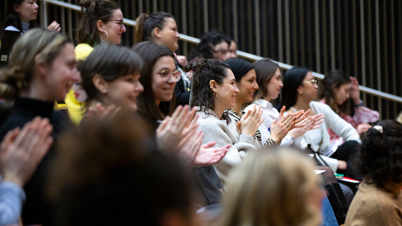Audience of diverse women sitting and clapping in a lecture hall or auditorium.