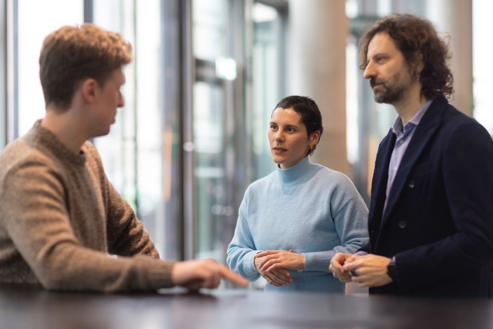 Three people having a focused conversation indoors near a window with a table in front of them.