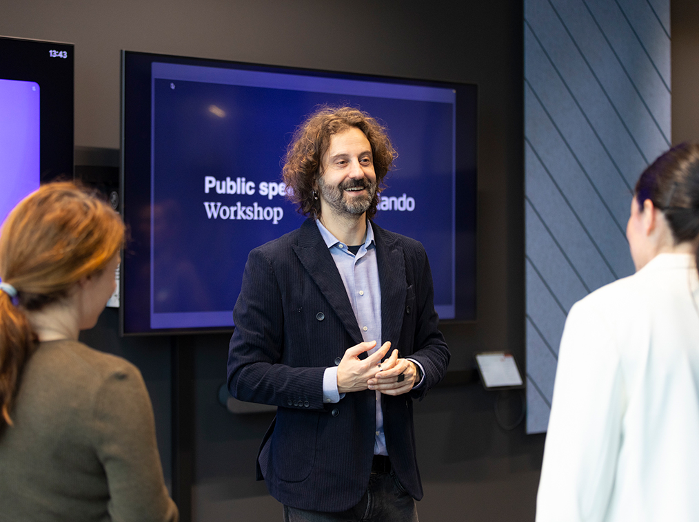 Man with curly hair and blazer leading a public speaking workshop to two attendees.