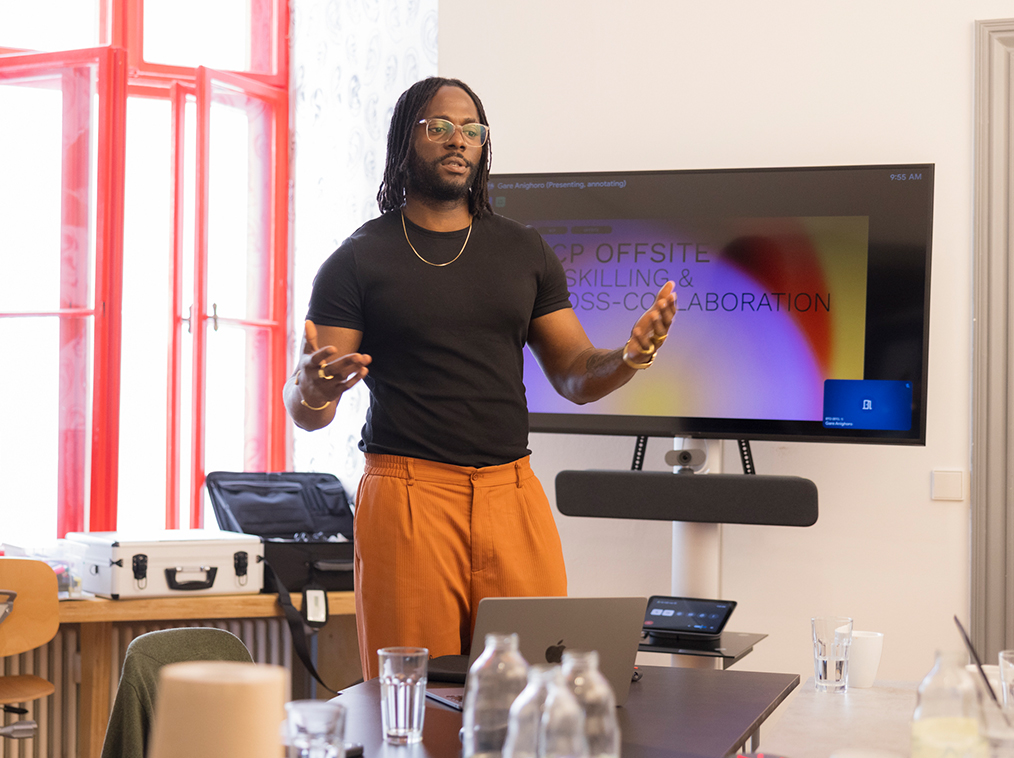 Man in black shirt and orange pants giving a presentation in a bright room with a screen behind him.