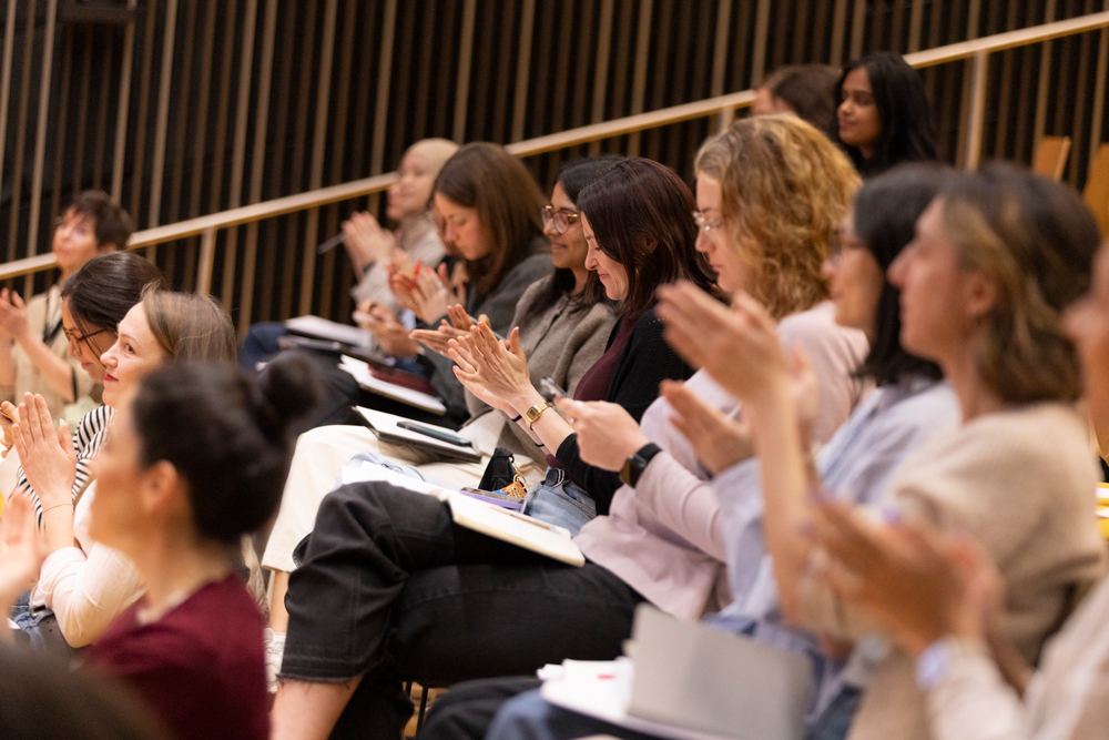 Diverse group of people seated in lecture hall clapping and smiling, some with notebooks.