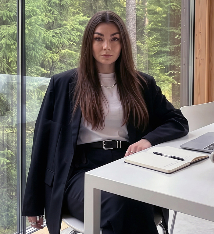 Woman with long brown hair in black suit sitting at a desk with notebook and laptop by window.