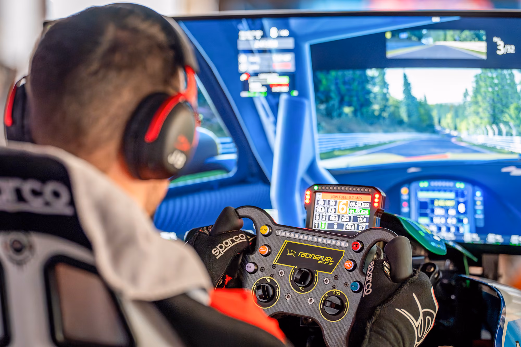 Nahaufnahme eines Fahrers mit Renn-Handschuhen am RacingFuel-Lenkrad, Strecke im Hintergrund auf dem Bildschirm