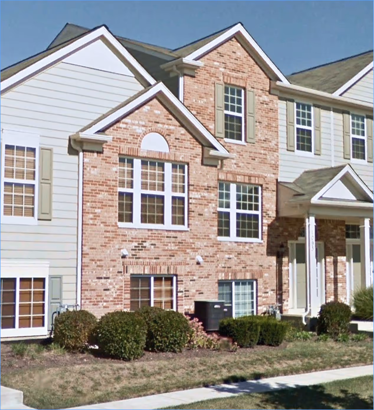 Townhouse building with brick and beige siding, multiple windows with white frames and green shutters, and neatly trimmed bushes in front.