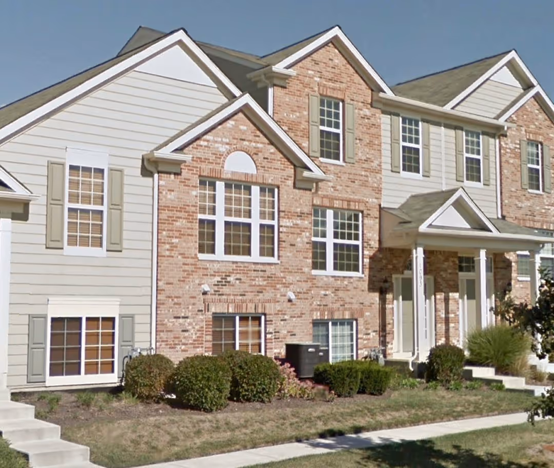 Row of two-story townhouses with brick and beige siding, featuring multiple windows, green shrubs, and a sidewalk in front.