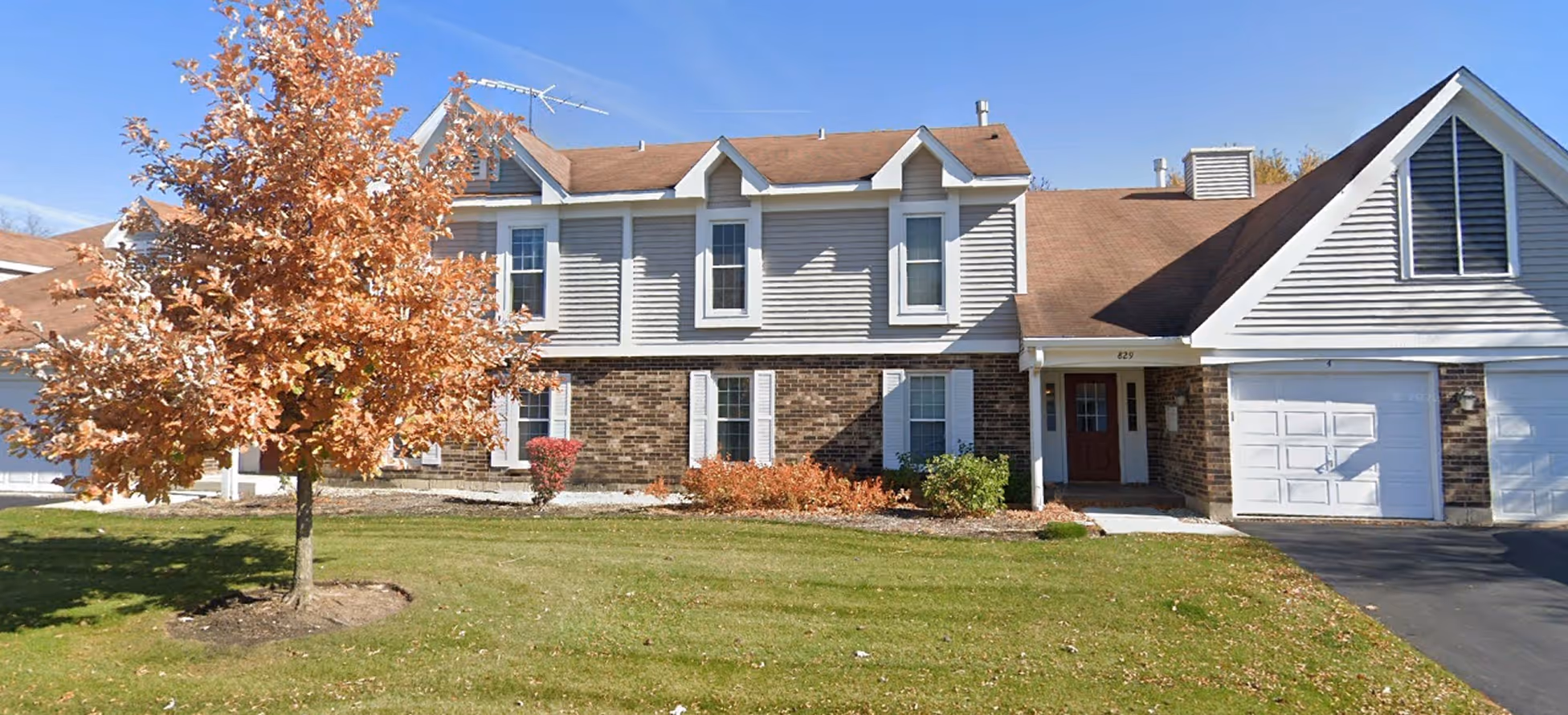 Two-story suburban house with brick and siding exterior, brown roof, single tree with autumn leaves, and attached garage.
