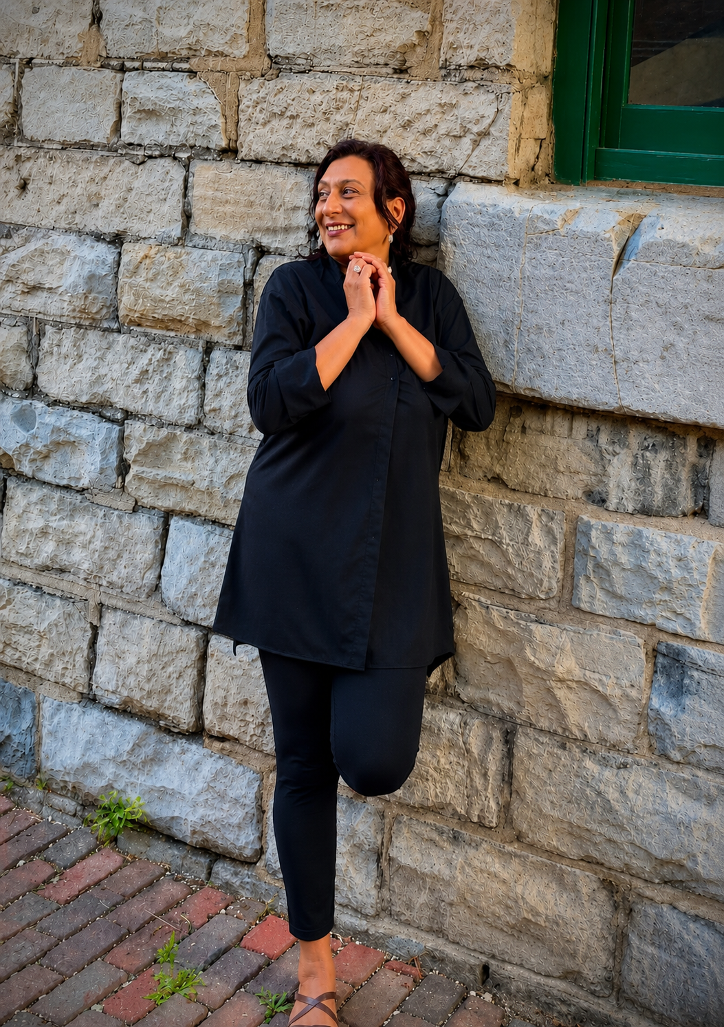 Smiling woman in black outfit leaning against textured stone wall with one knee raised.