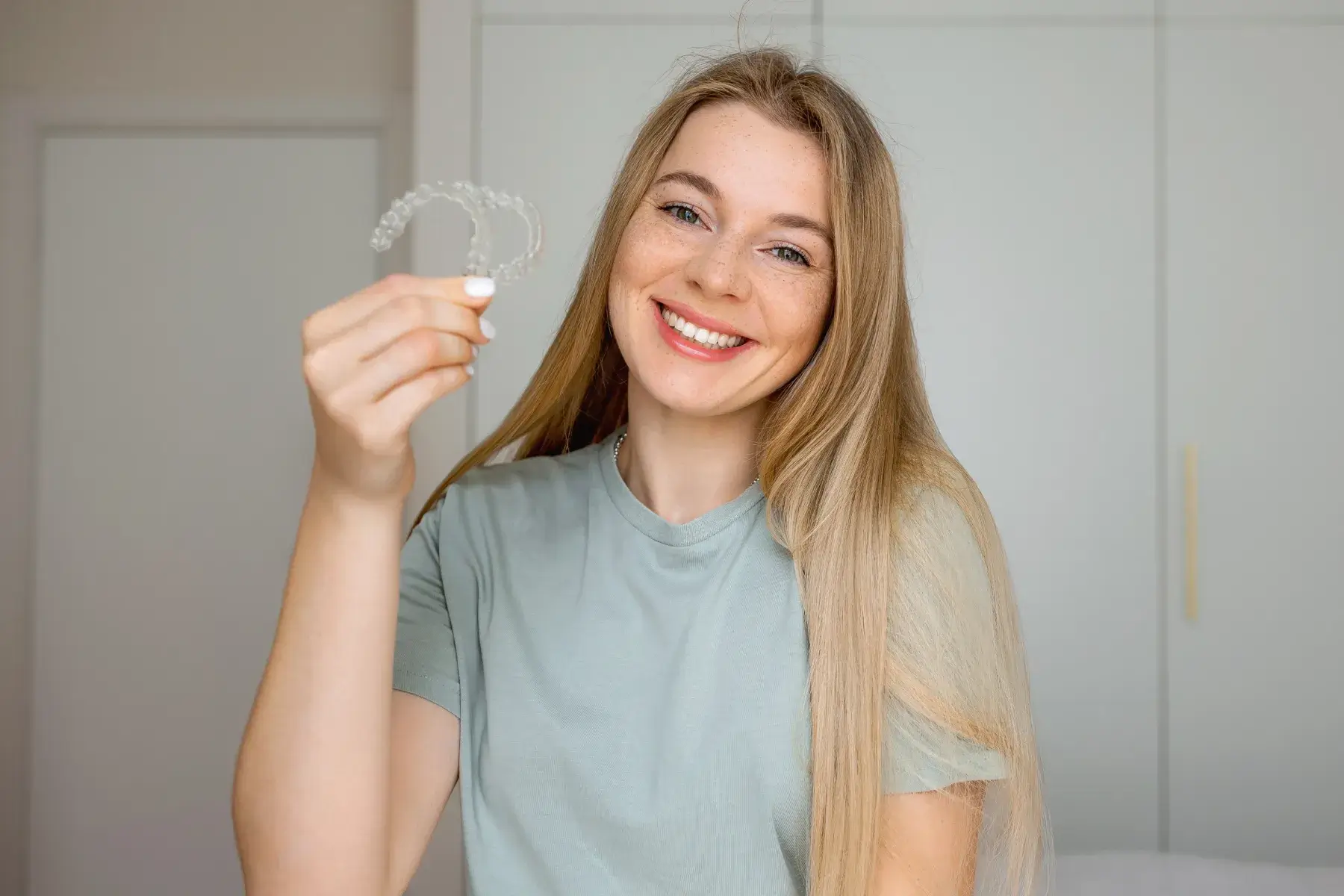 Smiling woman holding two clear dental aligners in her hand indoors.