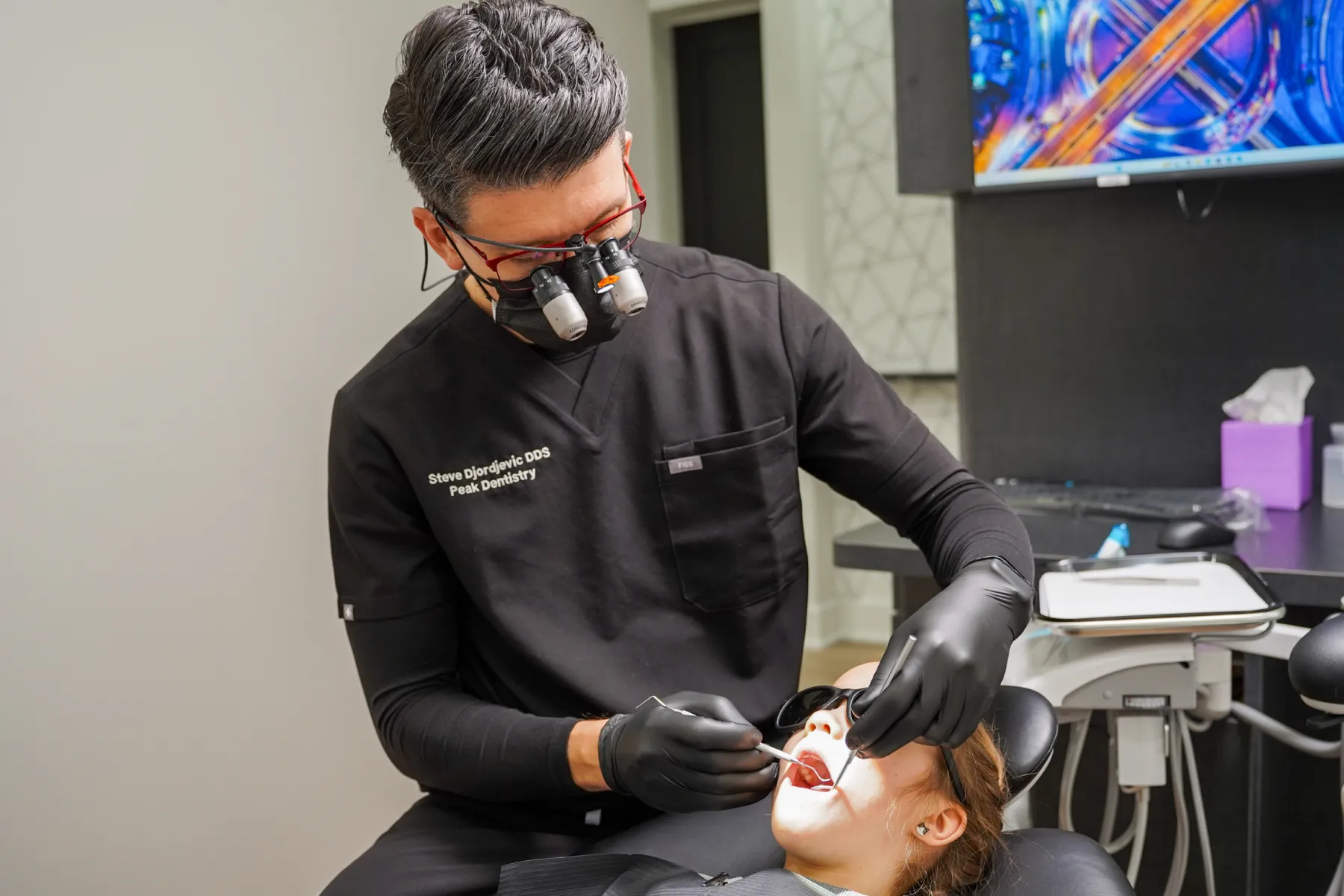 Dentist wearing magnifying loupes and black gloves examines a patient's mouth in a dental office.