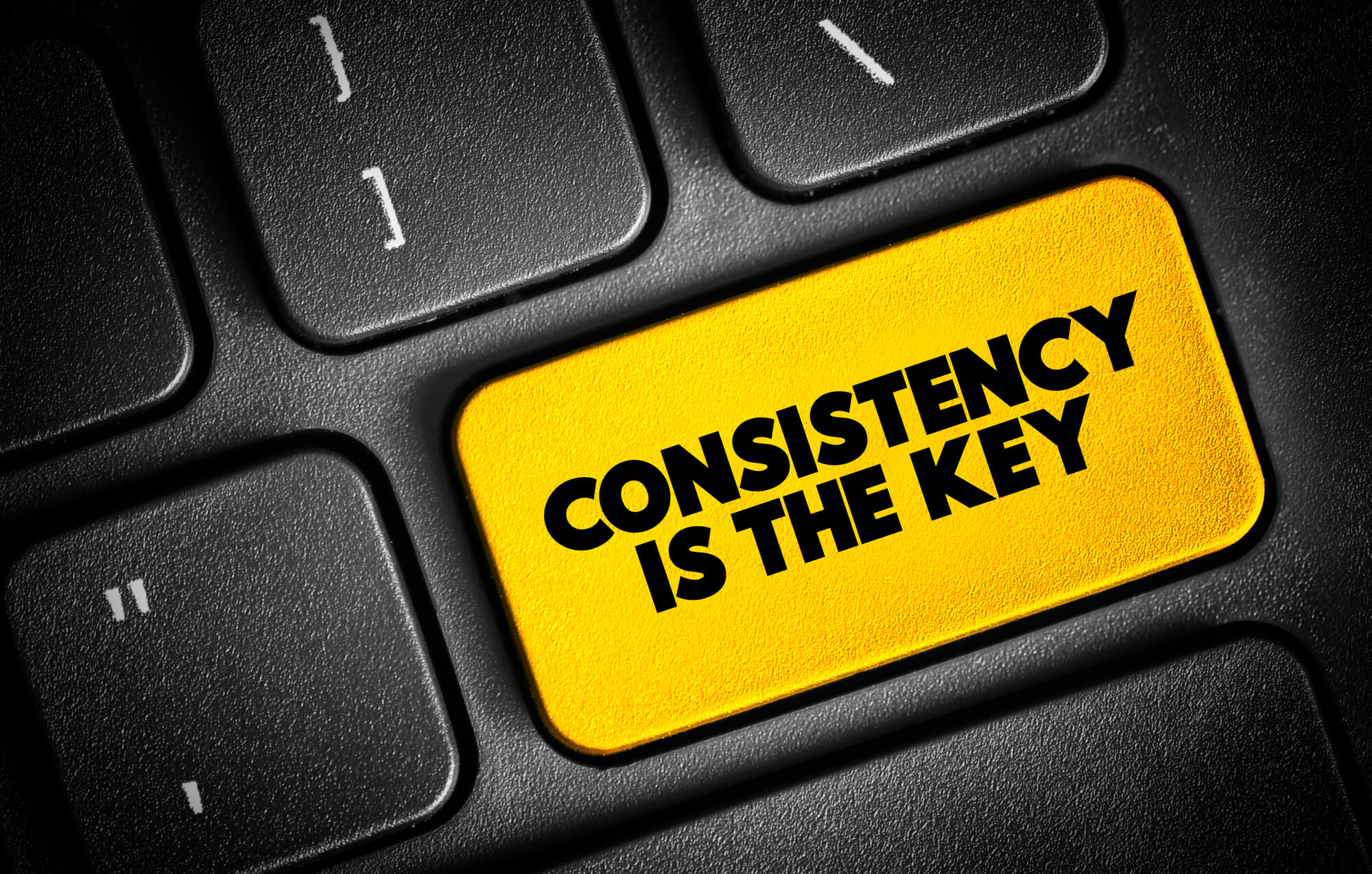 A black keyboard with a yellow key labeled "CONSISTENCY IS THE KEY" in bold black letters