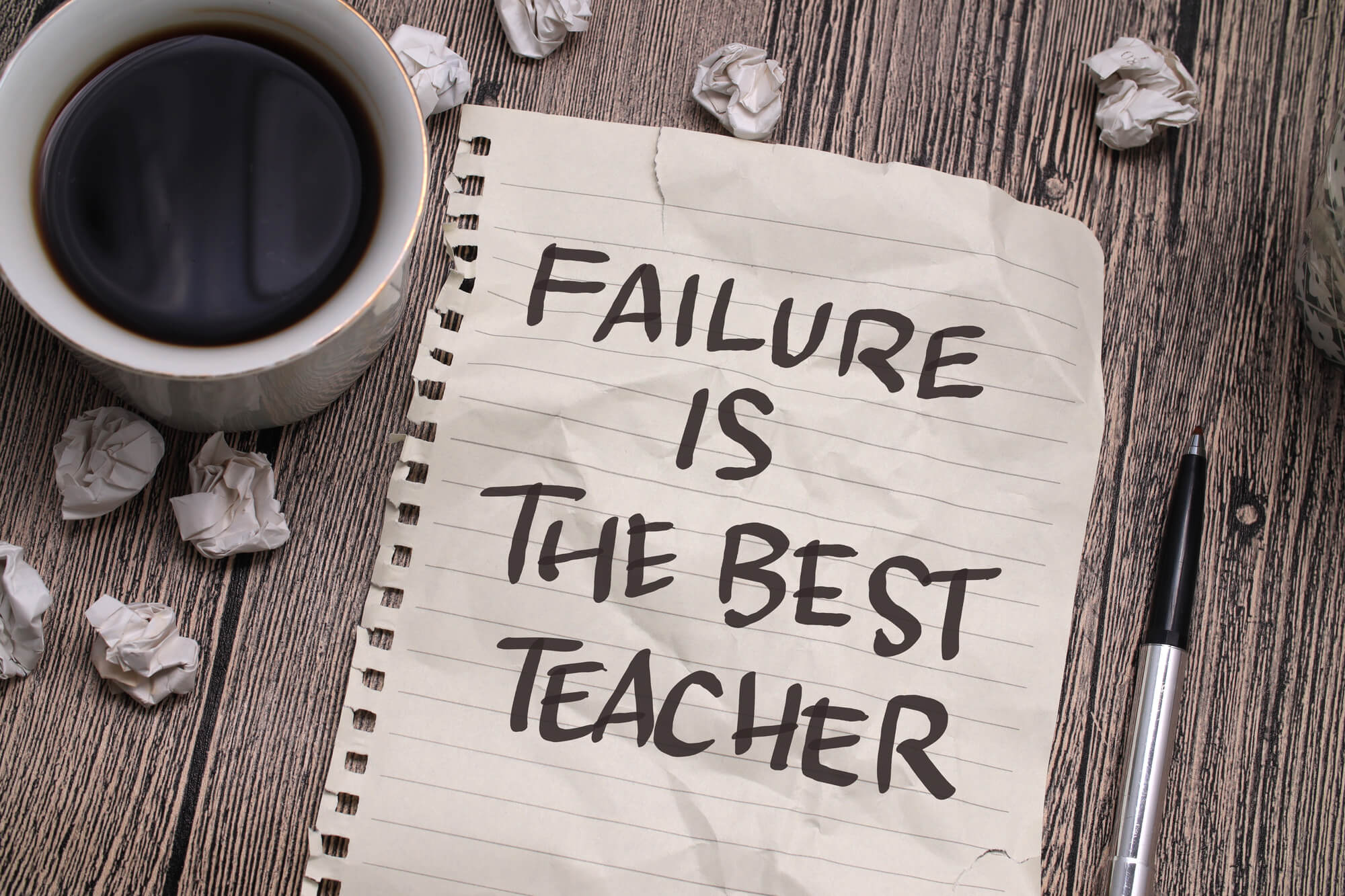 a cup of coffee, a pen, and crumpled paper surround a note that reads, "Failure is the best teacher"