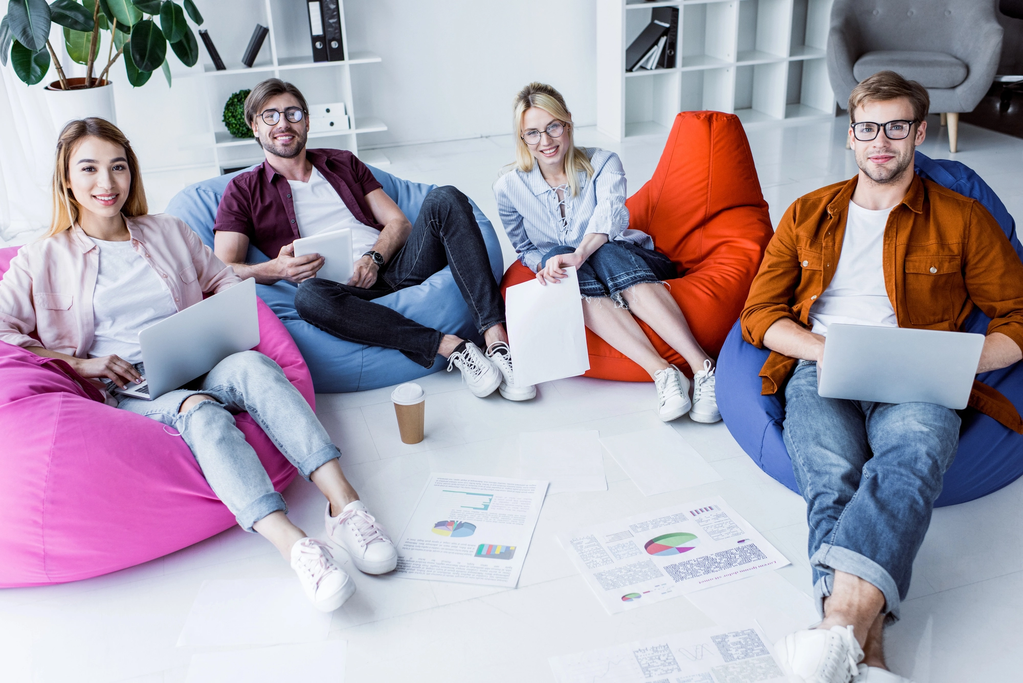 A small group of individuals sit on colorful bean bags, engaged with laptops and papers on the floor, in a modern workspace