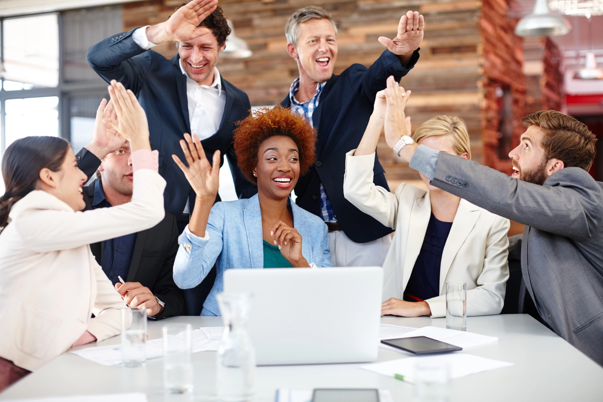 Group of diverse businesspeople around a table celebrating success with high-fives in a modern office setting