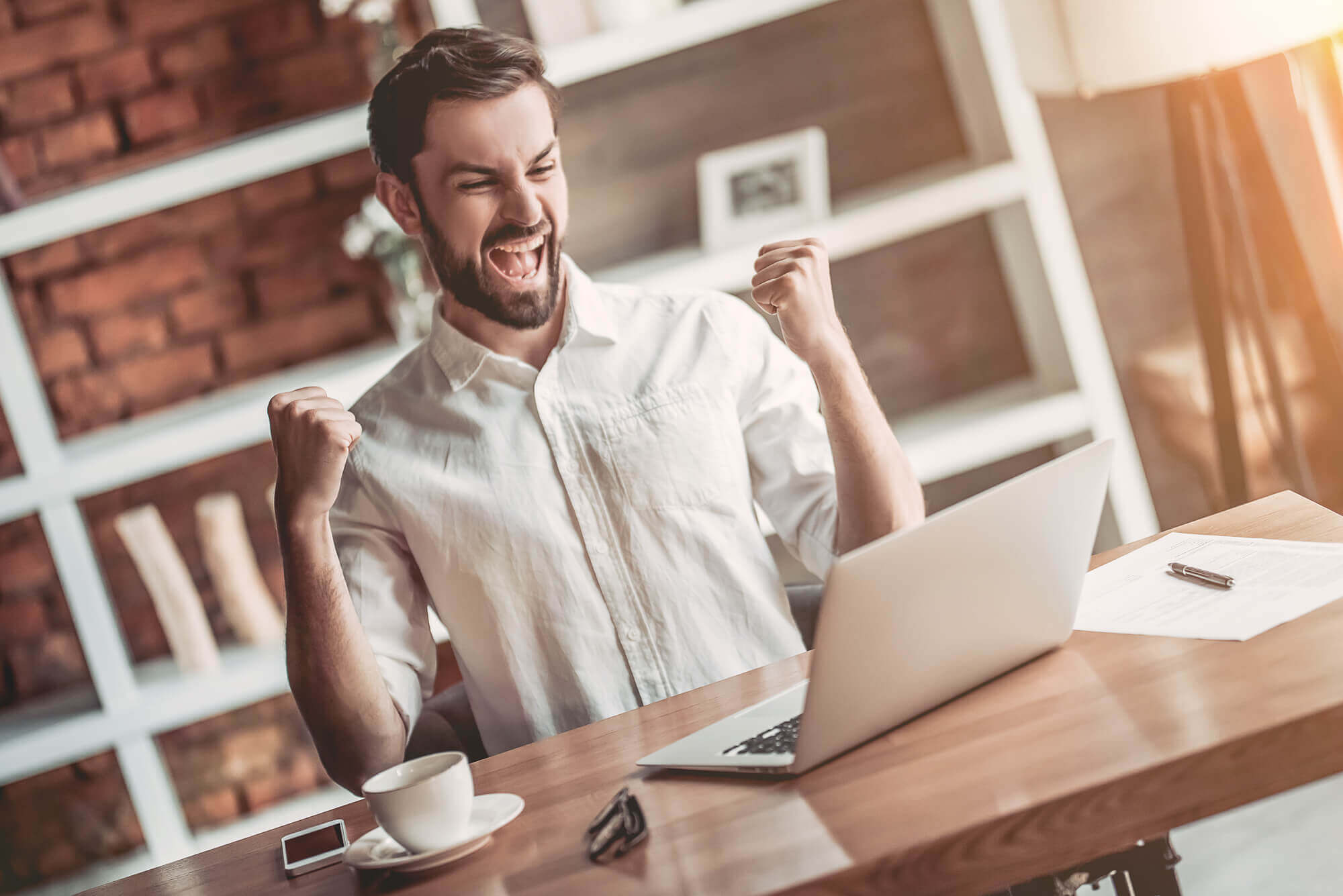 a young jubilant franchise owner at a home office desk raising fists in victory while looking at his laptop
