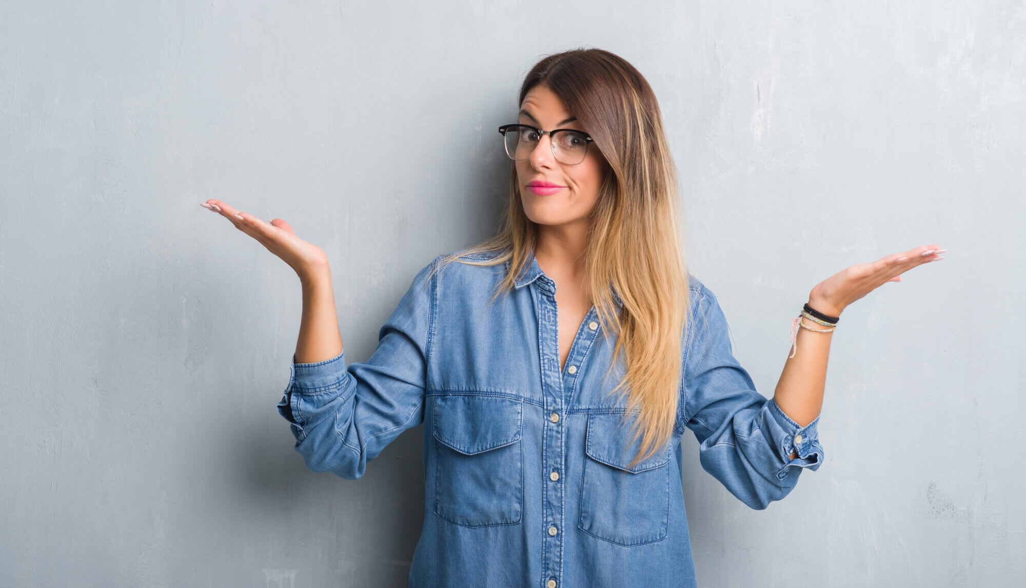 female marketer with glasses standing against a gray background shrugging with her palms up in a gesture of uncertainty
