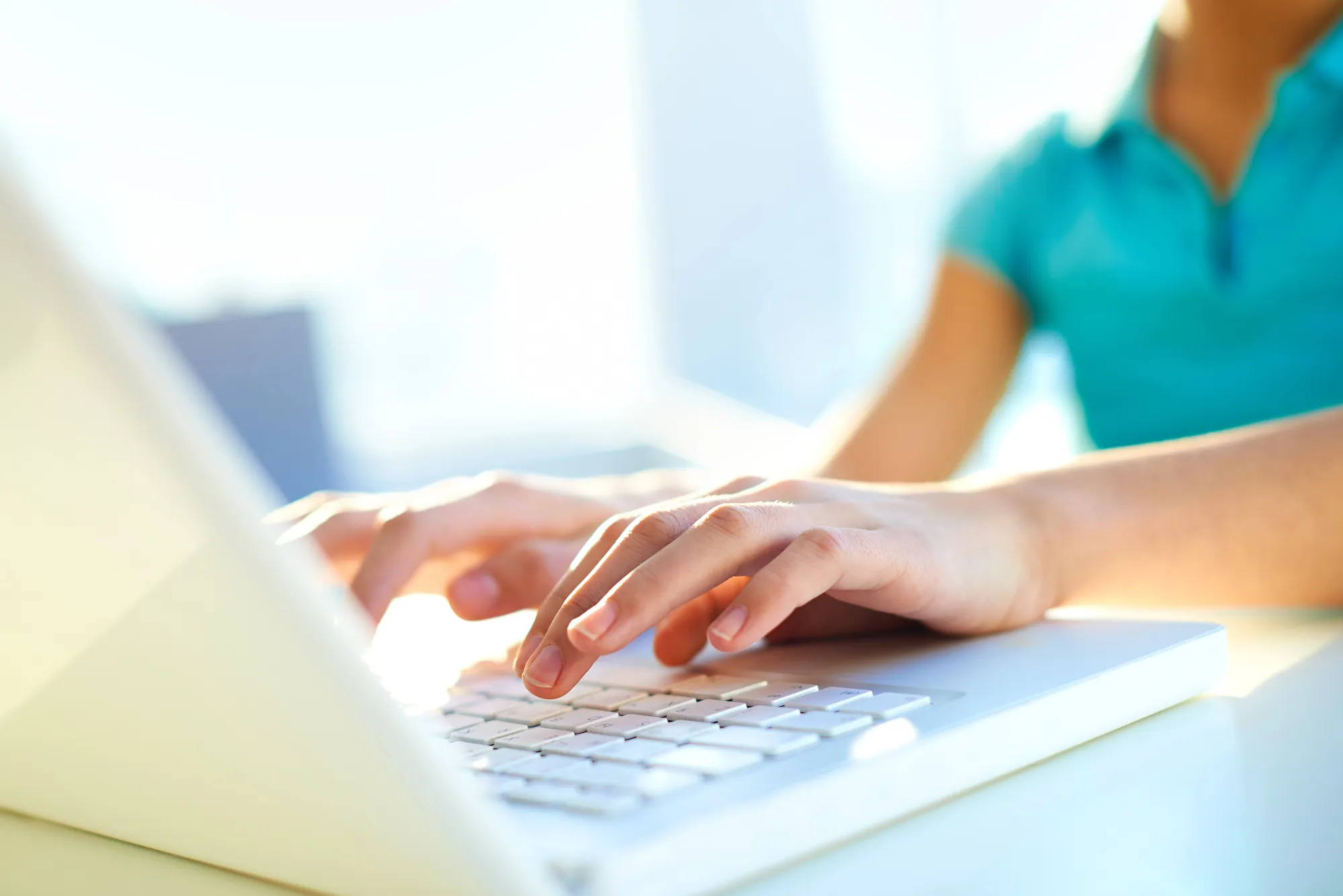 Close-up of hands typing on a white laptop keyboard in a bright sunlit office