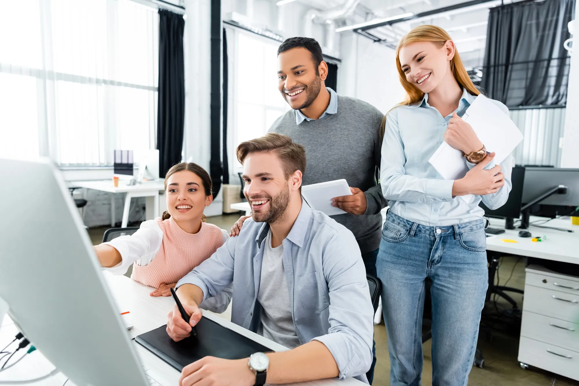 Four smiling coworkers gathered around a computer screen reviewing a website design in a modern office