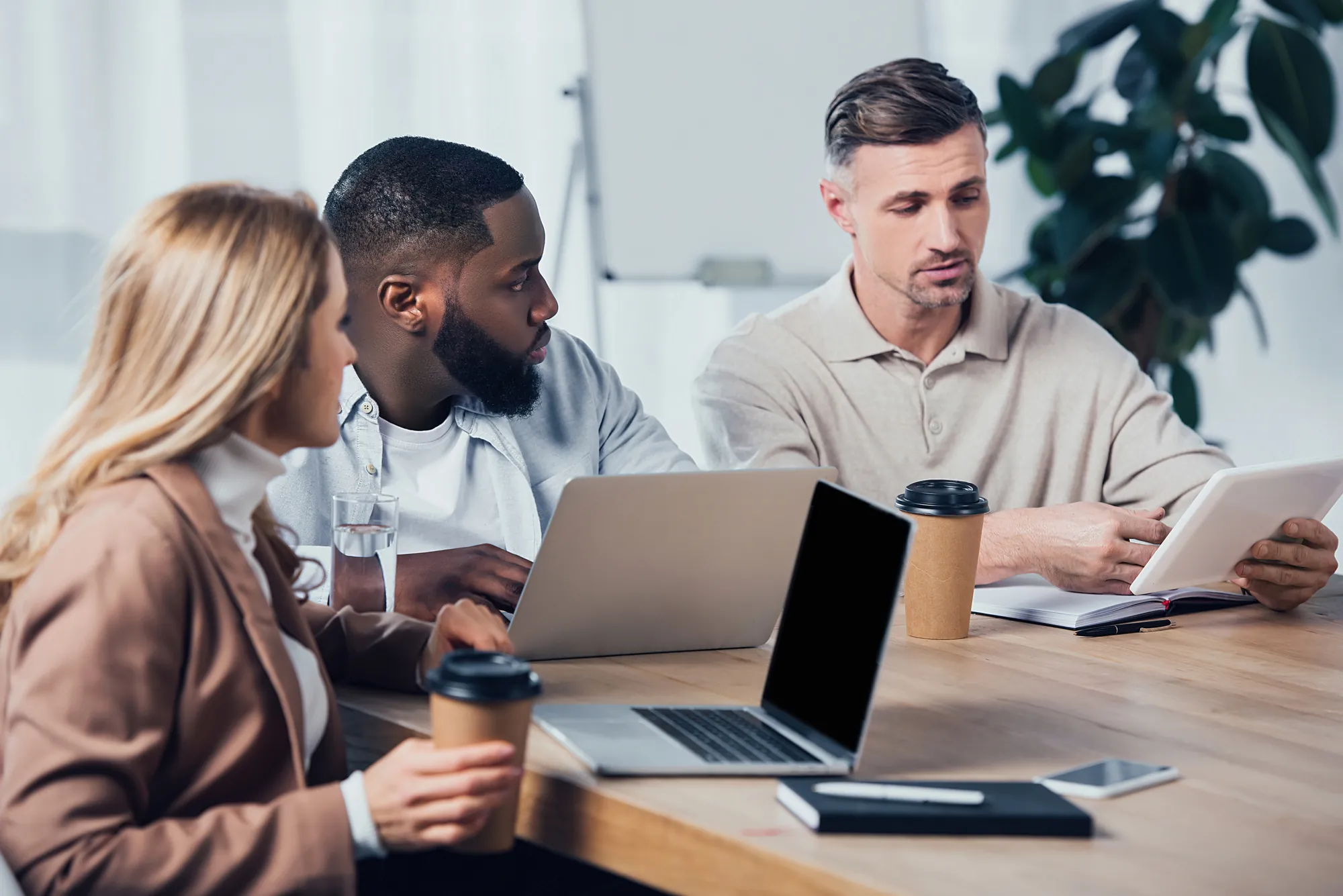 hree coworkers discussing work around laptops and coffee cups at a wooden table in a bright meeting room