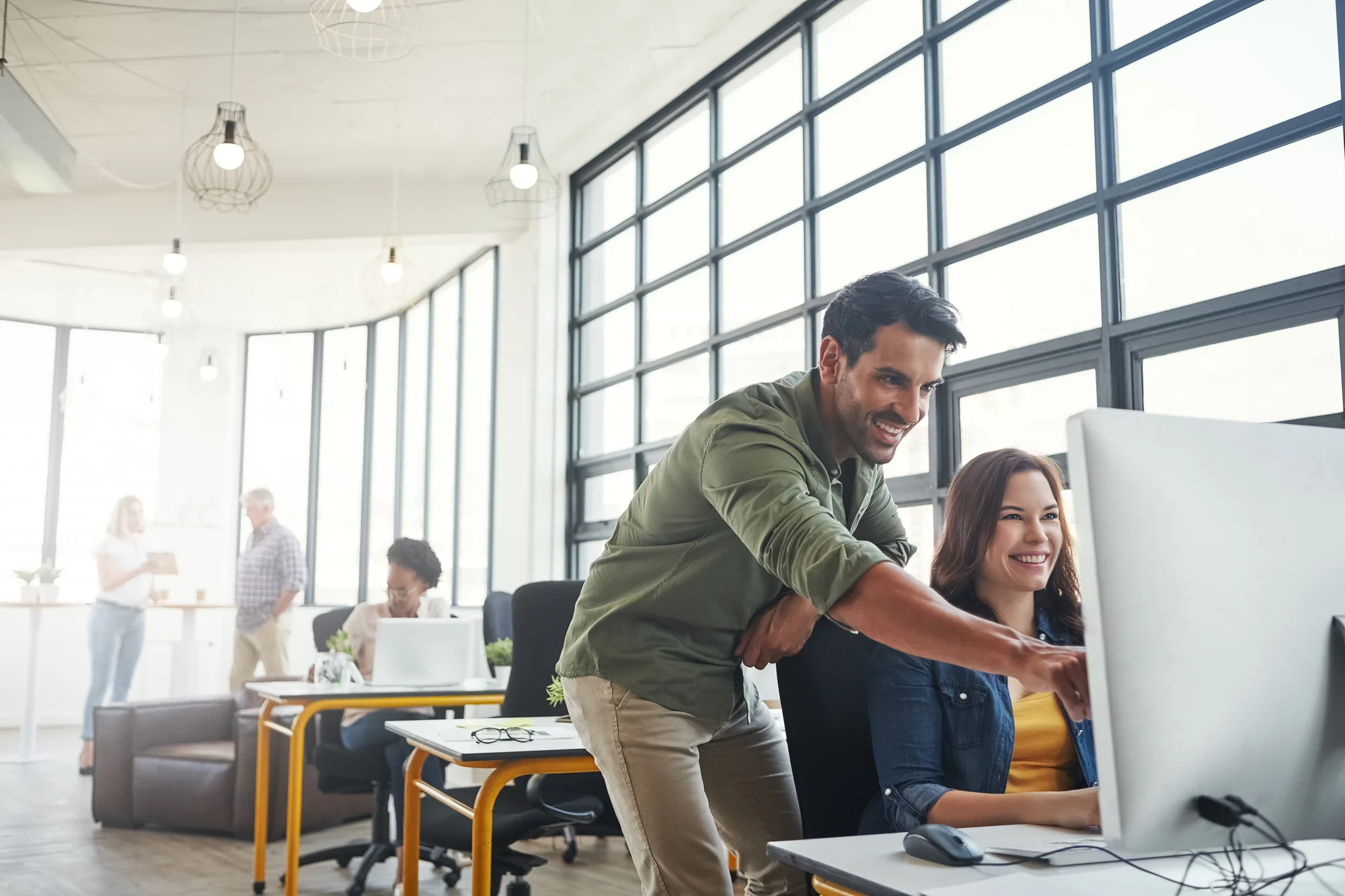Two coworkers smiling while pointing at a desktop monitor together in a bright open office