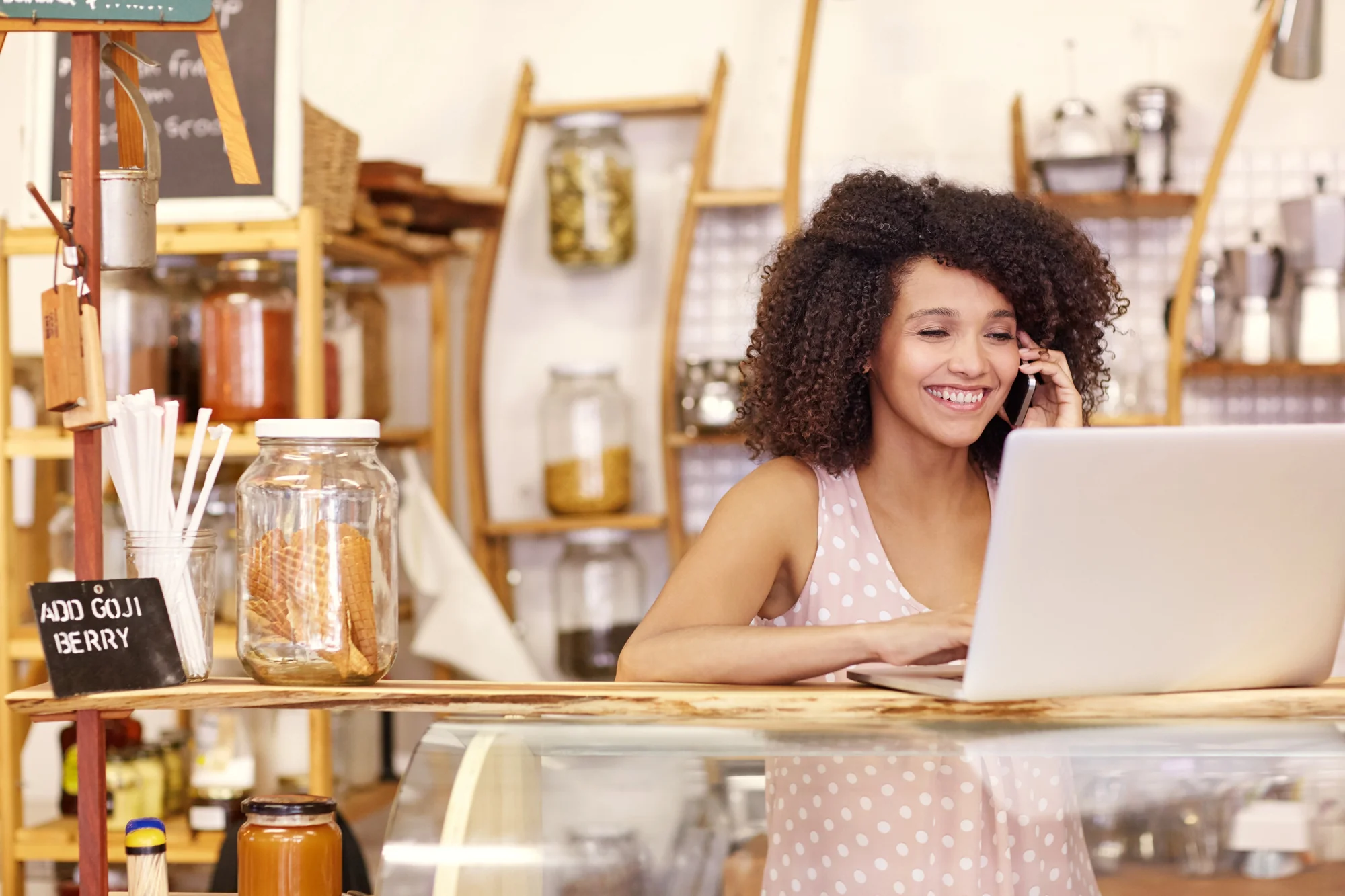 Smiling small business owner on the phone while working on a laptop at her shop counter, managing customer reviews with review generation services