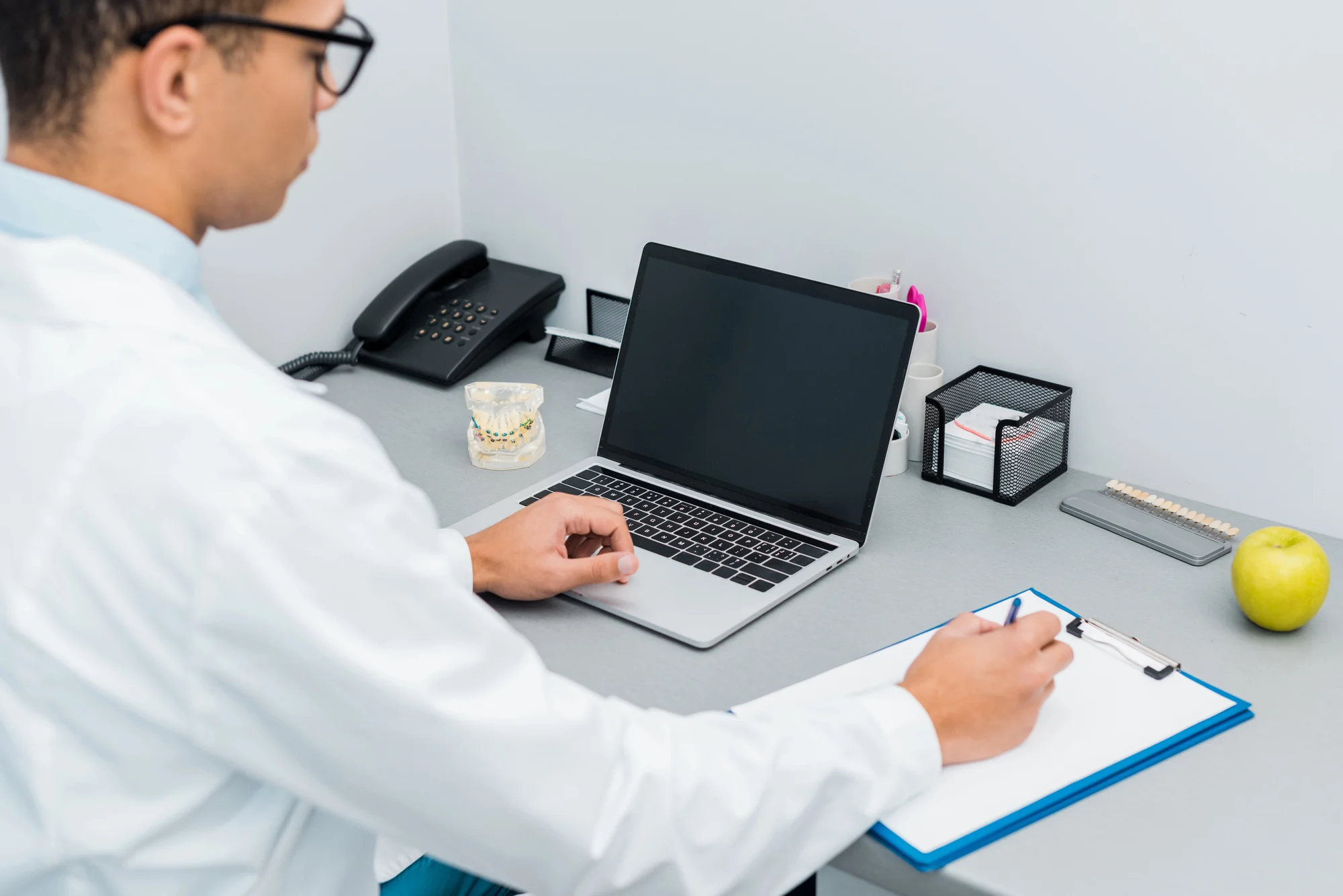 Dentist in a white coat at a desk with a laptop, clipboard, and dental model, managing the practice side of PPC for dentists