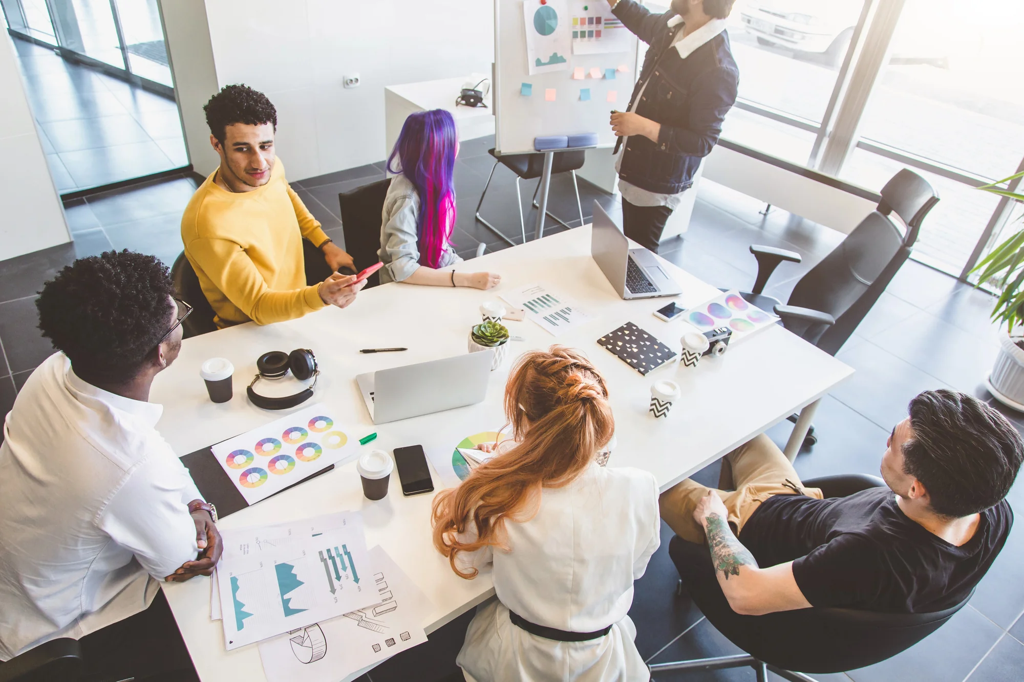Content team meeting around a table with charts and color swatches while a colleague presents ideas on a whiteboard